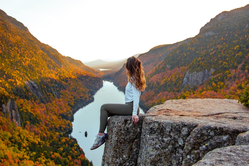 Woman sitting on a rocky cliff overlooking Lower Ausable Lake, surrounded by colorful fall foliage and layered Adirondack Mountains at golden hour